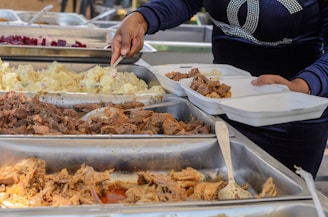 A person is serving food from buffet trays containing various dishes like meat, potatoes, and another food item into divided takeaway containers. The person is using a serving utensil and is wearing a dark blue long-sleeved shirt.
