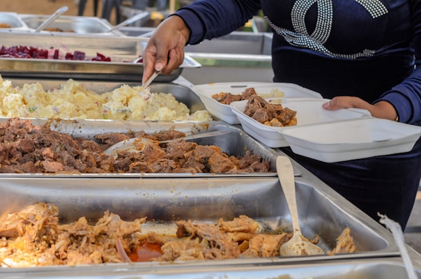 A person is serving food from buffet trays containing various dishes like meat, potatoes, and another food item into divided takeaway containers. The person is using a serving utensil and is wearing a dark blue long-sleeved shirt.
