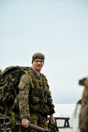 A person dressed in full military gear stands outdoors, holding equipment and wearing a camouflage uniform. The background suggests a clear or lightly cloudy sky.