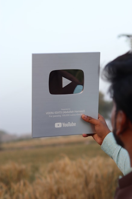 A person holds a YouTube Silver Play Button award, which is given for achieving 100,000 subscribers. The background features a field under a clear sky.
