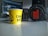 A yellow paper cup with the words 'Chai Kafi' printed on it is placed on a table next to a pair of black JBL headphones. The background is slightly blurred, suggesting an indoor setting, possibly an office or studio environment.