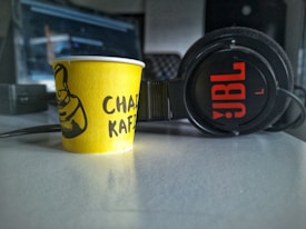 A yellow paper cup with the words 'Chai Kafi' printed on it is placed on a table next to a pair of black JBL headphones. The background is slightly blurred, suggesting an indoor setting, possibly an office or studio environment.
