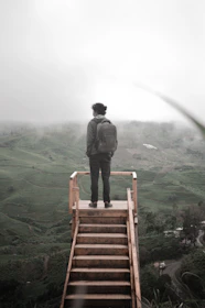 A person wearing a backpack stands at the top of a wooden staircase, overlooking a vast, misty landscape of green hills. The scene appears to be serene and reflective, with low clouds and fog covering parts of the background.