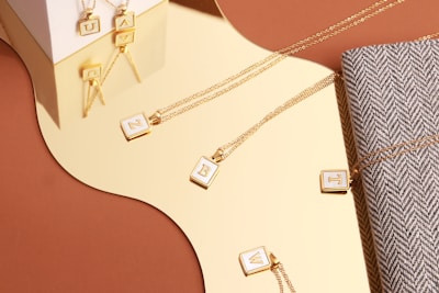 Gold necklaces with square pendants featuring individual letters are arranged on a shiny reflective surface. Some pendants rest directly on a herringbone fabric, adding texture to the composition. The setting includes a mix of geometric shapes and natural curves, contrasting with the linear chains and structured pendants. Lighting highlights the metallic sheen and casts soft reflections.