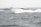 A sleek white yacht navigates through choppy waters, with waves crashing against its hull. In the background, a smaller, blue utility vessel is visible, contributing to the sense of activity at sea. The sky is overcast, adding to the energetic and slightly dramatic scene.