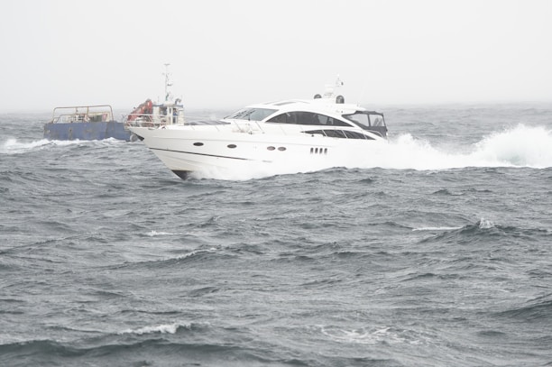 A sleek white yacht navigates through choppy waters, with waves crashing against its hull. In the background, a smaller, blue utility vessel is visible, contributing to the sense of activity at sea. The sky is overcast, adding to the energetic and slightly dramatic scene.