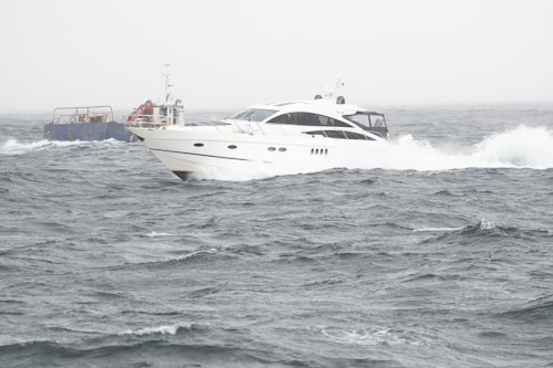 A sleek white yacht navigates through choppy waters, with waves crashing against its hull. In the background, a smaller, blue utility vessel is visible, contributing to the sense of activity at sea. The sky is overcast, adding to the energetic and slightly dramatic scene.