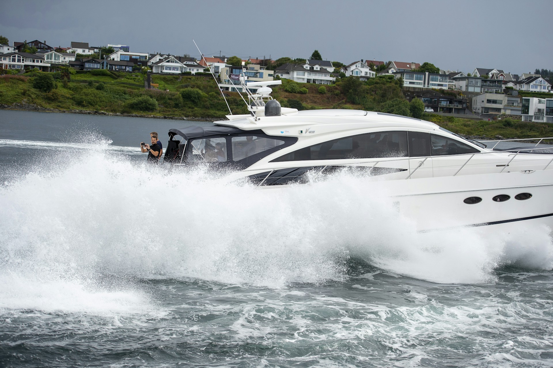 A white yacht creating a large splash as it moves swiftly through the water. In the background, there is a shoreline with several houses and greenery. A person is visible on the deck of the yacht, appearing to be taking photos or videos.