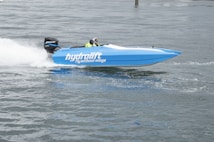 A blue speedboat with the words 'hydrolift fly without wings' printed on its side is racing across the water, creating a trail of white spray behind it. The driver is wearing a yellow and black outfit, and the boat is equipped with a Mercury outboard motor.