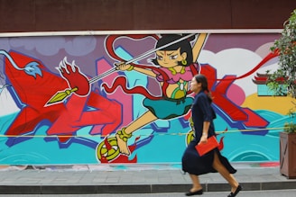 A colorful mural featuring a stylized animated character wielding a spear, set against a vivid background with abstract shapes and a mythological theme. A woman in a blue dress is walking in front of the mural, carrying a red clutch.