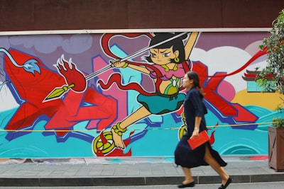 A colorful mural featuring a stylized animated character wielding a spear, set against a vivid background with abstract shapes and a mythological theme. A woman in a blue dress is walking in front of the mural, carrying a red clutch.