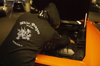 A person wearing a black hoodie with a 'White Dragon Sports Gear' logo on the back is working on the engine of an orange car with an open hood. The scene is in a dimly lit parking garage.