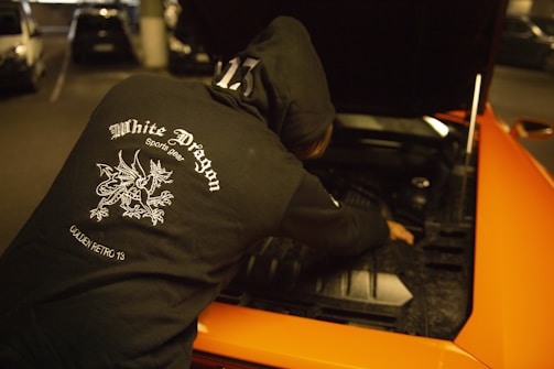 A person wearing a black hoodie with a 'White Dragon Sports Gear' logo on the back is working on the engine of an orange car with an open hood. The scene is in a dimly lit parking garage.