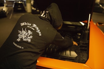 A person wearing a black hoodie with a 'White Dragon Sports Gear' logo on the back is working on the engine of an orange car with an open hood. The scene is in a dimly lit parking garage.