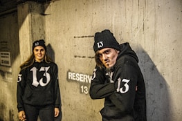 A man and a woman are wearing matching black hoodies and beanies with the number 13 printed on them. They are standing against a concrete wall with the word 'RESERVED' and the number 13 painted on it. The man on the right appears to have a serious or intense expression, while the woman on the left is smiling.