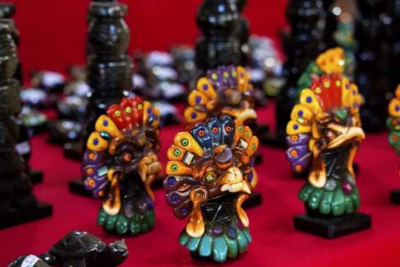 A collection of colorful, intricately designed ornamental figures resembling stylized creatures, arranged on a bright red surface. The figures feature vibrant patterns with a mix of colors including blue, green, orange, and red, and are positioned in several rows.