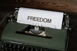 A vintage typewriter sits on a wooden surface with a sheet of paper emerging from it. The word 'FREEDOM' is typed in bold, capital letters on the paper.