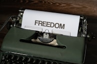 A vintage typewriter sits on a wooden surface with a sheet of paper emerging from it. The word 'FREEDOM' is typed in bold, capital letters on the paper.
