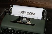 A vintage typewriter sits on a wooden surface with a sheet of paper emerging from it. The word 'FREEDOM' is typed in bold, capital letters on the paper.