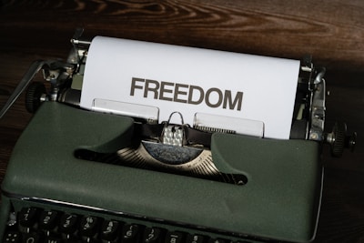 A vintage typewriter sits on a wooden surface with a sheet of paper emerging from it. The word 'FREEDOM' is typed in bold, capital letters on the paper.