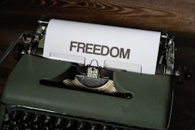 A vintage typewriter sits on a wooden surface with a sheet of paper emerging from it. The word 'FREEDOM' is typed in bold, capital letters on the paper.