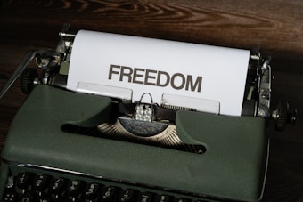 A vintage typewriter sits on a wooden surface with a sheet of paper emerging from it. The word 'FREEDOM' is typed in bold, capital letters on the paper.