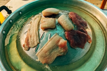 A metal grill pan placed over a heat source with several pieces of raw meat and fat cooking, likely used for a barbecue or grilling session.