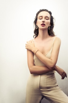 A woman with shoulder-length hair and closed eyes wearing a sleeveless top and trousers, adorned with earrings and a bracelet. Her arms are crossed gently over her chest, conveying a serene expression against a neutral background.