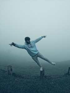 A person is balancing on a post amid a foggy landscape, with arms outstretched and one leg extended behind them. The scene is set in a misty environment that conveys a sense of calm and tranquility.