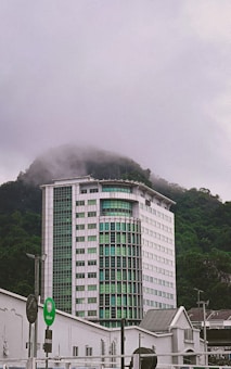 A tall modern building with a facade of green glass windows stands amidst fog, partially covering the lush green forested hill behind it. In the foreground, several signs, including one with the word 'Keluar', are visible alongside smaller structures.