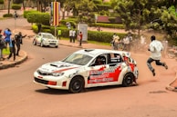 A white rally car with vibrant red and black decals is navigating a sharp corner on a city road. The car has 'International University of East Africa' written on its side. A man is seen jumping away from the road as if startled by the car's sudden approach. Spectators, some holding cameras, are gathered on the roadside capturing the action. Another car is visible in the background around the curve. The setting appears to be a lively urban environment with well-maintained greenery and pathways.
