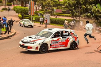 A white rally car with vibrant red and black decals is navigating a sharp corner on a city road. The car has 'International University of East Africa' written on its side. A man is seen jumping away from the road as if startled by the car's sudden approach. Spectators, some holding cameras, are gathered on the roadside capturing the action. Another car is visible in the background around the curve. The setting appears to be a lively urban environment with well-maintained greenery and pathways.