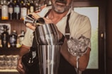 A bartender is preparing a cocktail using a shaker and a jigger. The background displays a variety of liquor bottles on shelves, suggesting a bar setting. Ice-filled glassware is visible, indicating a focus on mixology.