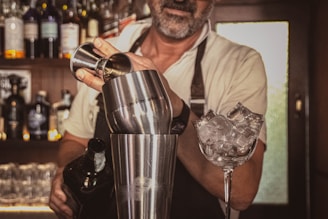 A bartender is preparing a cocktail using a shaker and a jigger. The background displays a variety of liquor bottles on shelves, suggesting a bar setting. Ice-filled glassware is visible, indicating a focus on mixology.