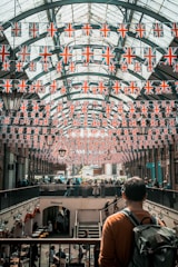A marketplace with an arched glass ceiling is decorated with numerous British flags. People are walking and shopping in the busy space below, where cafes and shops are visible. A man stands in the foreground, looking over the scene with a backpack on his shoulder.