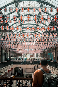 A marketplace with an arched glass ceiling is decorated with numerous British flags. People are walking and shopping in the busy space below, where cafes and shops are visible. A man stands in the foreground, looking over the scene with a backpack on his shoulder.
