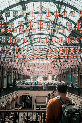 A marketplace with an arched glass ceiling is decorated with numerous British flags. People are walking and shopping in the busy space below, where cafes and shops are visible. A man stands in the foreground, looking over the scene with a backpack on his shoulder.