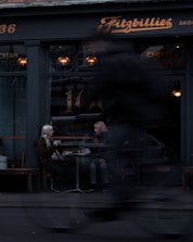 Two people sit at a small table outside a cafe called Fitzbillies, engaged in conversation. The seating area is near a large window with warm lighting inside. A passing cyclist is blurred in the foreground, indicating motion and urban life.