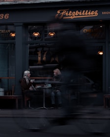 Two people sit at a small table outside a cafe called Fitzbillies, engaged in conversation. The seating area is near a large window with warm lighting inside. A passing cyclist is blurred in the foreground, indicating motion and urban life.
