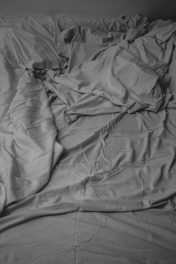 Crinkled and unkempt bedsheets are spread across a bed surface, creating a pattern of folds and creases. The monochromatic color scheme suggests a simple and minimalistic setting.