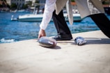 A person is kneeling on a dock beside a body of water, handling fish that lie on the ground. A boat is visible in the background, while the person’s attire blends formal elements with practical footwear suitable for work.