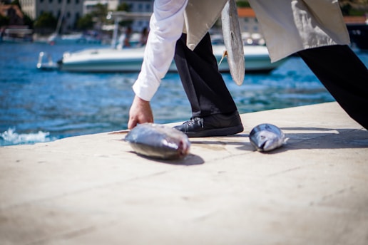 A person is kneeling on a dock beside a body of water, handling fish that lie on the ground. A boat is visible in the background, while the person’s attire blends formal elements with practical footwear suitable for work.