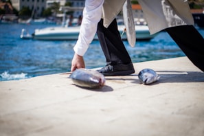 A person is kneeling on a dock beside a body of water, handling fish that lie on the ground. A boat is visible in the background, while the person’s attire blends formal elements with practical footwear suitable for work.