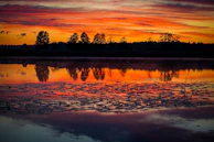 A vibrant sunset over a quiet lake, reflecting fiery oranges and purples in the still water.