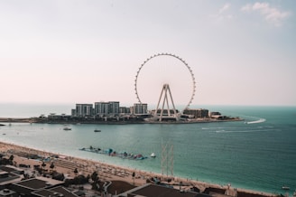 A large Ferris wheel stands prominently on a small island surrounded by calm blue waters. Nearby, modern buildings and a bustling waterfront with beach umbrellas line the shore. Several small boats and jet skis move across the water.