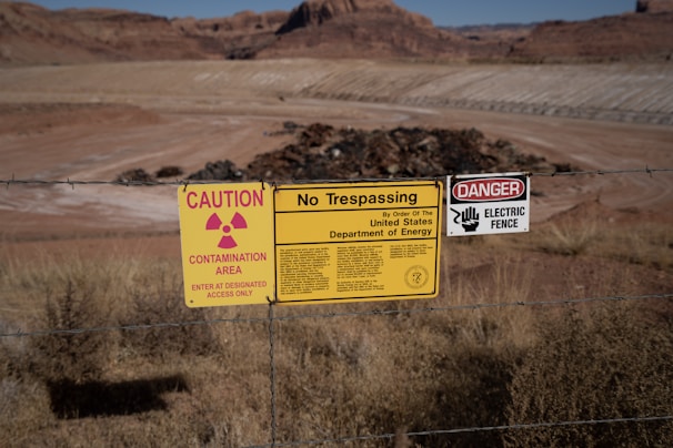 A chain-link fence marked by multiple warning signs indicates a restricted area. The signs include warnings for radiation contamination, trespassing, and an electric fence, attributed to the United States Department of Energy. The background shows a barren landscape with rocky terrain and dry grass.