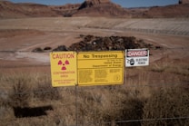 A chain-link fence marked by multiple warning signs indicates a restricted area. The signs include warnings for radiation contamination, trespassing, and an electric fence, attributed to the United States Department of Energy. The background shows a barren landscape with rocky terrain and dry grass.