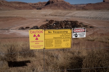 A chain-link fence marked by multiple warning signs indicates a restricted area. The signs include warnings for radiation contamination, trespassing, and an electric fence, attributed to the United States Department of Energy. The background shows a barren landscape with rocky terrain and dry grass.