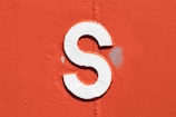 A bold white letter 'S' is painted on a textured, bright orange surface. The letter appears slightly raised, casting a subtle shadow on the background. There is a visible patch of lighter discoloration near the letter.