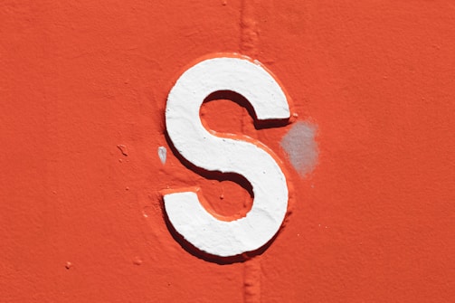 A bold white letter 'S' is painted on a textured, bright orange surface. The letter appears slightly raised, casting a subtle shadow on the background. There is a visible patch of lighter discoloration near the letter.